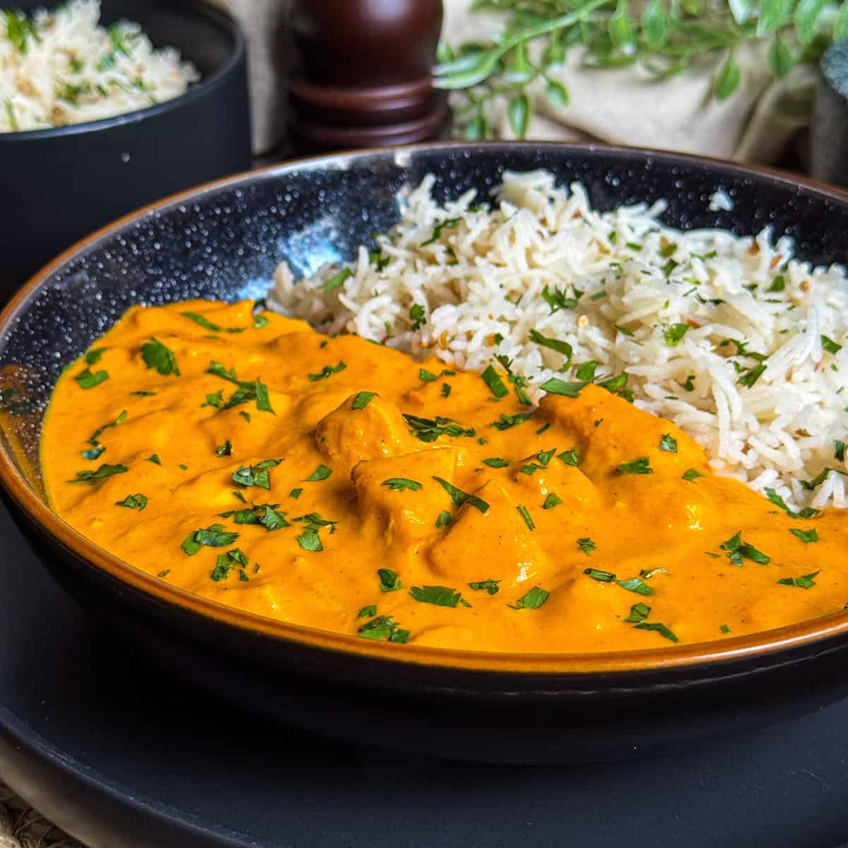 A black bowl filled with creamy orange curry garnished with chopped herbs, served alongside fluffy white rice. The dish is set on a dark plate with blurred background elements.