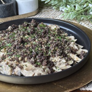A black plate holds creamy penne pasta topped with finely crumbled ground meat and chopped herbs, set on a gold charger with greenery and jars in the background.