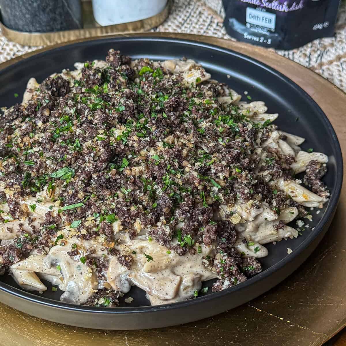 A black plate filled with creamy pasta topped with a generous layer of finely crumbled black pudding and garnished with chopped parsley, set on a gold charger.