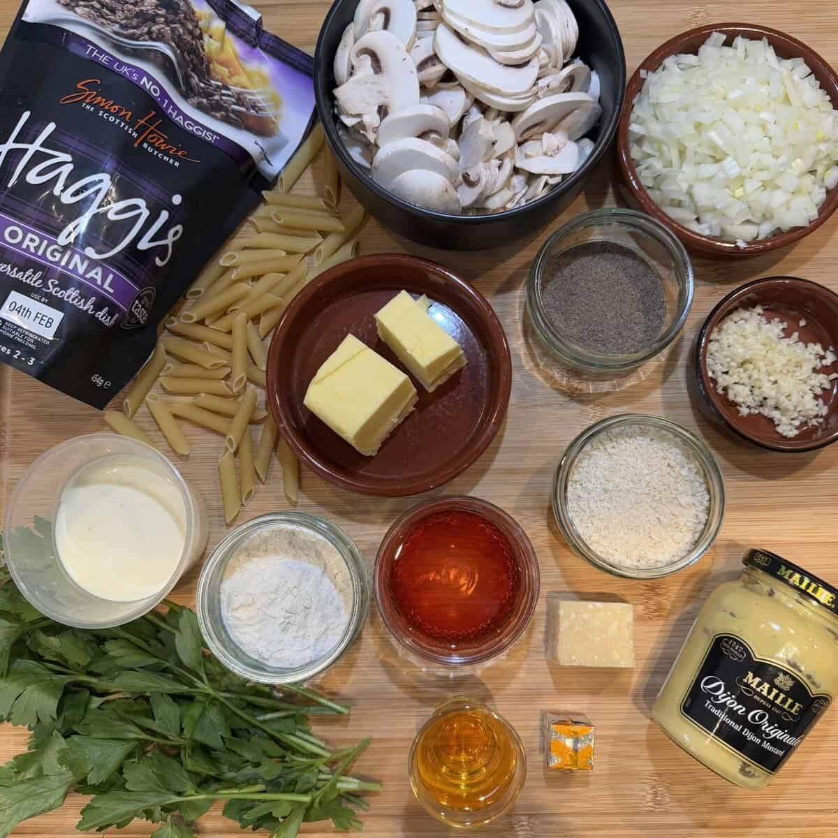 A wooden surface displays ingredients for a recipe: sliced mushrooms, diced onions, butter, penne pasta, flour, breadcrumbs, jars of spices, chopped parsley, mustard, whisky, cheese, and a packet of haggis.