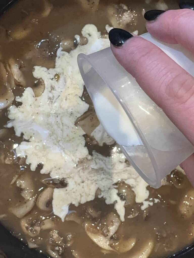A hand with black-painted nails pours cream from a small cup into a pot of soup with mushrooms, meat, and pasta.