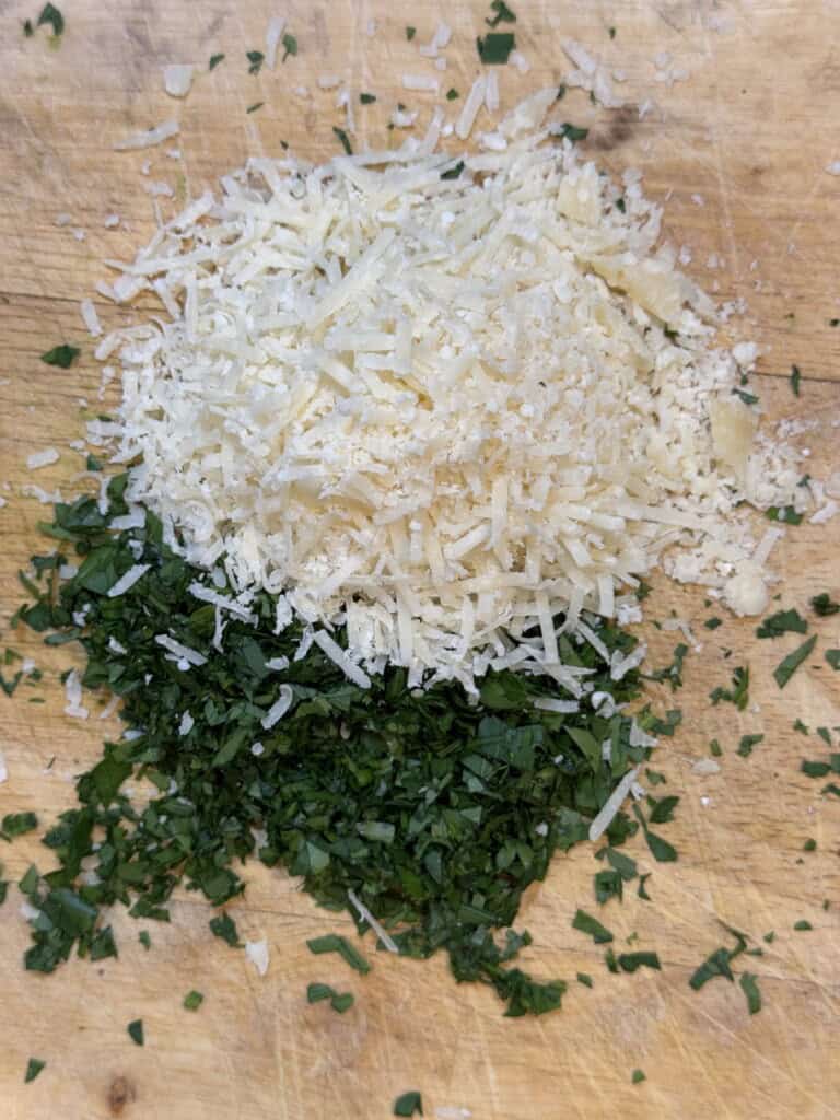 A pile of grated Parmesan cheese sits next to a small pile of chopped fresh parsley on a wooden cutting board.