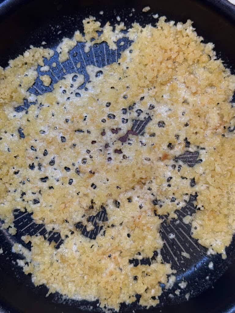 Golden breadcrumbs are being toasted in melted butter in a black nonstick pan. The breadcrumbs are spread unevenly, showing some browning and sizzling across the surface.