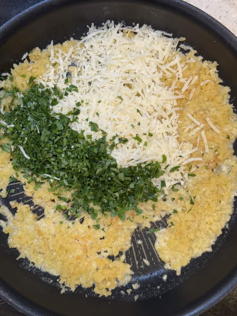 Chopped parsley and shredded cheese are added to sautéed minced garlic in a frying pan.