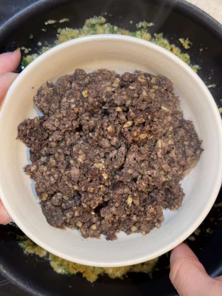 A person holds a white bowl filled with a dark, crumbly mixture over a pan containing chopped vegetables.