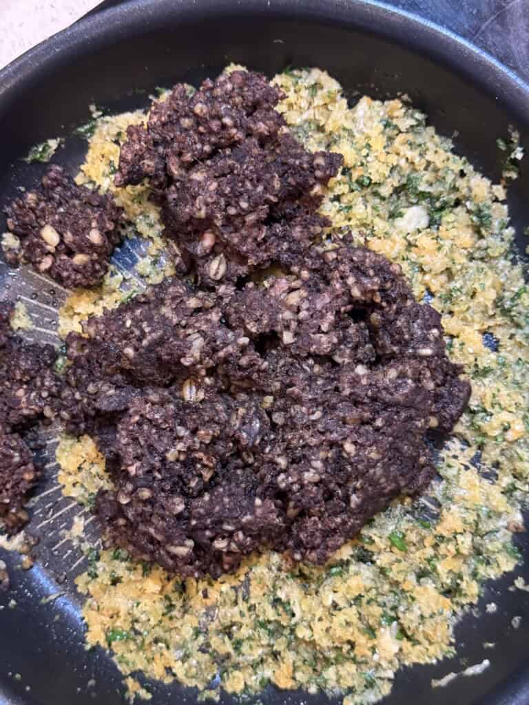 A close-up of a pan containing a mixture of finely chopped green herbs and yellow crumbs, with several dark brown, crumbly, cooked patties or chunks on top.