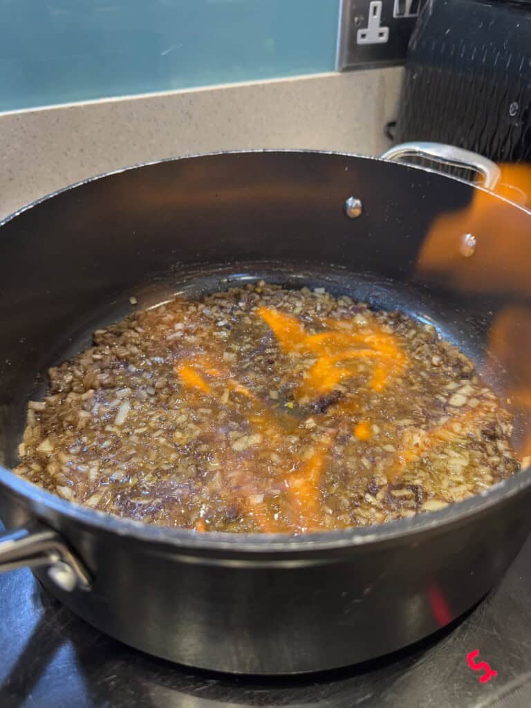 Chopped onions are sautéing in a large black saucepan, with orange flames visible, indicating the onions are being flambéed on a stovetop. A toaster and power outlets are in the background.