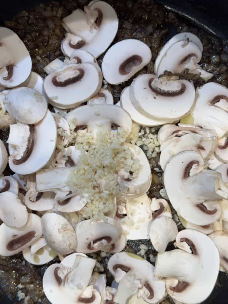 Sliced white mushrooms and minced garlic cooking in a pan with sautéed onions.