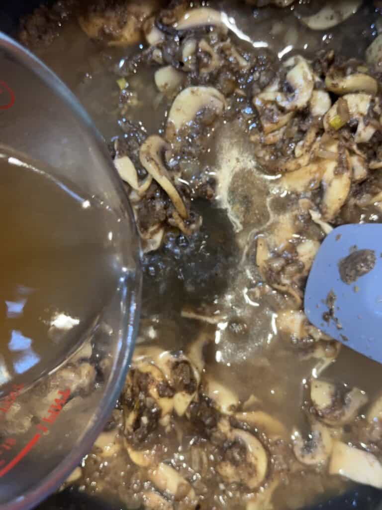 Close-up of sliced mushrooms being cooked in a pan with liquid being poured in from a measuring cup and a spatula stirring the mixture.