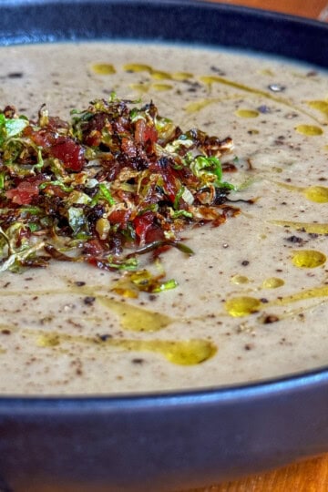 A bowl of creamy Brussel Sprout soup topped with a garnish of chopped bacon and shredded brussel sprouts, drizzled with olive oil, and served on a rustic wooden table.