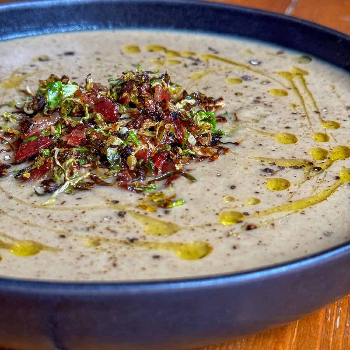 A bowl of creamy Brussel Sprout soup topped with a garnish of chopped bacon and shredded brussel sprouts, drizzled with olive oil, and served on a rustic wooden table.