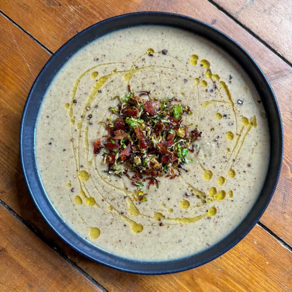 A bowl of creamy Brussel Sprout soup topped with a garnish of chopped bacon and shredded brussel sprouts, drizzled with olive oil, and served on a rustic wooden table.