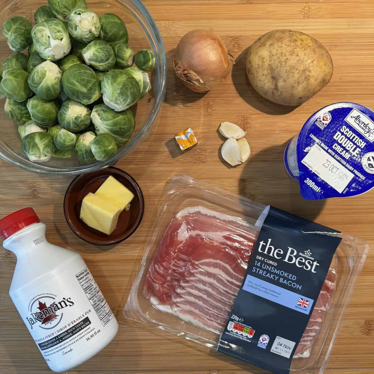A top-down view of ingredients on a wooden surface: brussels sprouts, onion, potato, double cream, bacon, butter, milk, garlic cloves, and a stock cube.