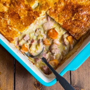 A close-up of a baked casserole dish with golden-brown pastry crust, filled with creamy stew containing chunks of chicken, ham and sliced carrots. A serving spoon is placed in the dish, revealing the filling.