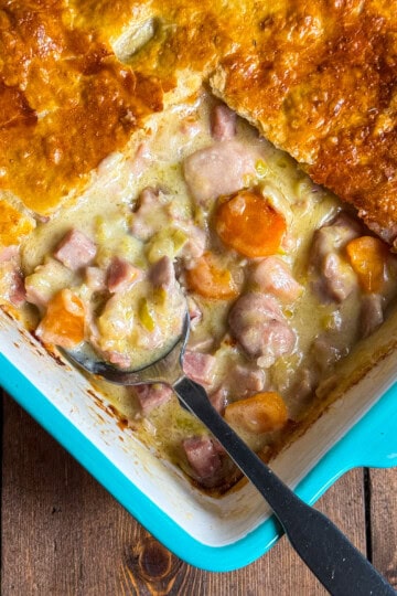 A close-up of a baked casserole dish with golden-brown pastry crust, filled with creamy stew containing chunks of chicken, ham and sliced carrots. A serving spoon is placed in the dish, revealing the filling.