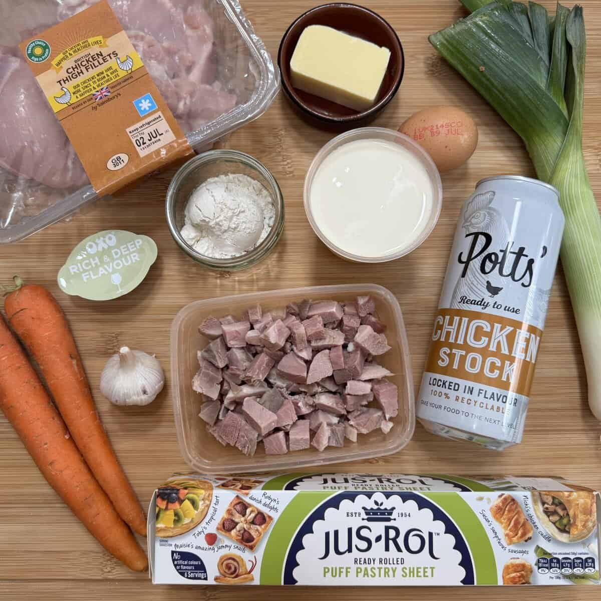 Ingredients for a homemade pie arranged on a wooden surface, including chicken thighs, diced ham, carrots, leek, garlic, flour, cream, an egg, chicken stock, rich beef stock pot, and a roll of puff pastry.