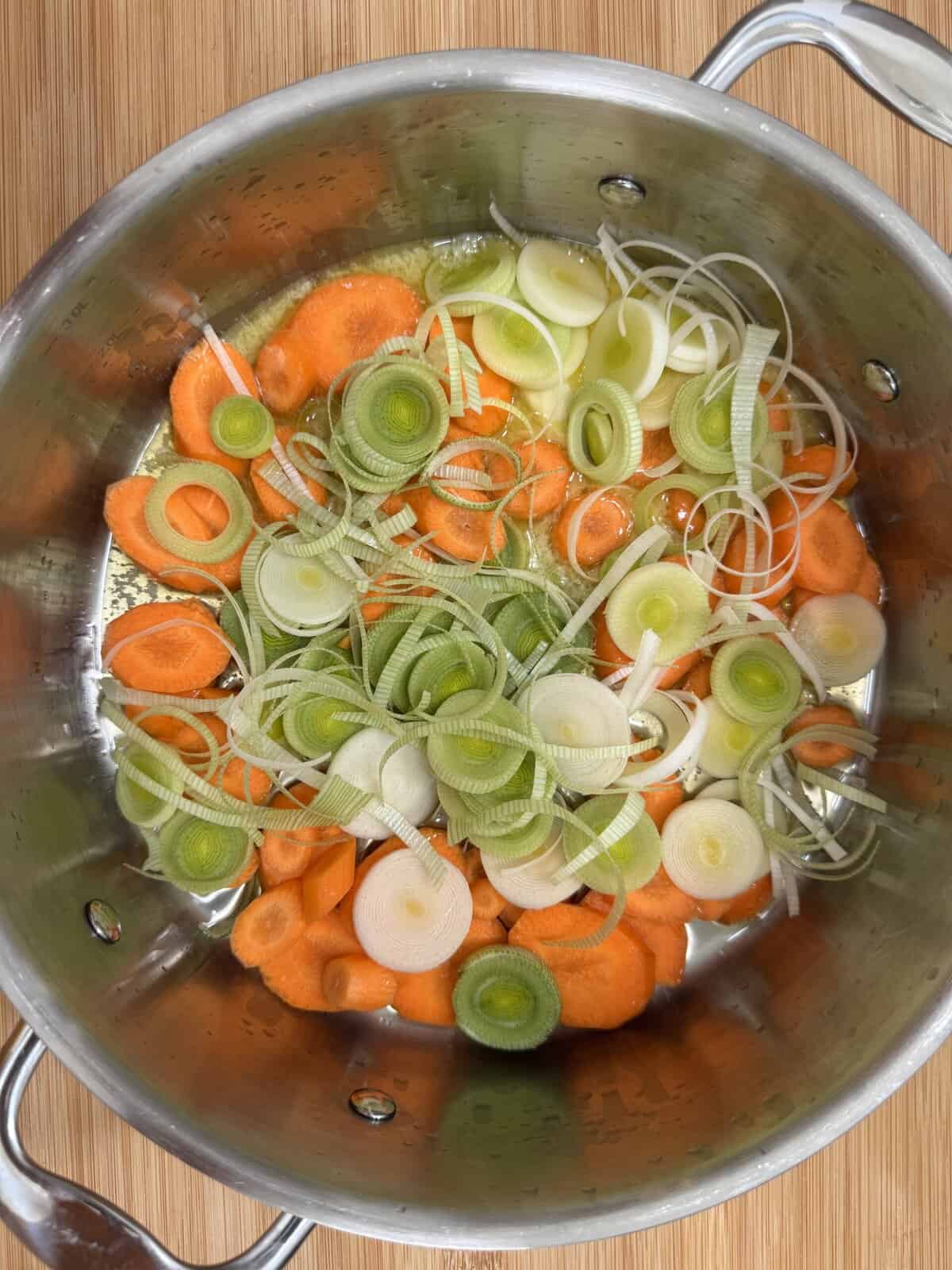 Sliced carrots and leeks in a stainless steel pot on a wooden surface, ready to be cooked.
