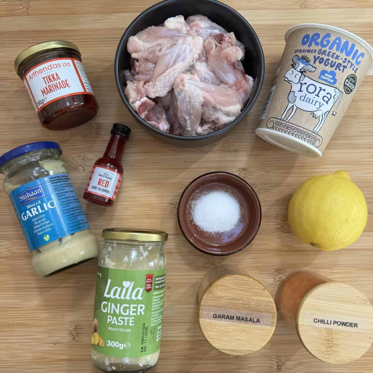 Ingredients for making chicken tikka placed on a wooden surface: raw chicken wings, Greek yogurt, tikka marinade, garlic paste, ginger paste, red food coloring, salt, lemon, garam masala, and chili powder.