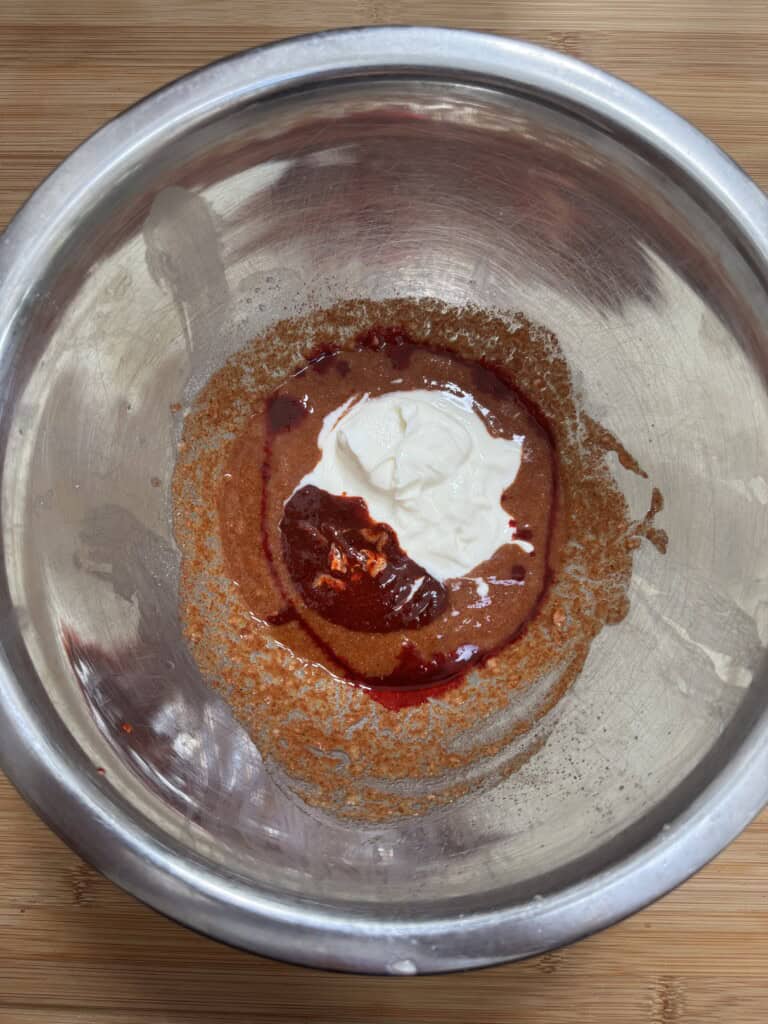 A metal mixing bowl with a thick brown batter, a dollop of white cream, and a swirl of red liquid on top, placed on a wooden surface.