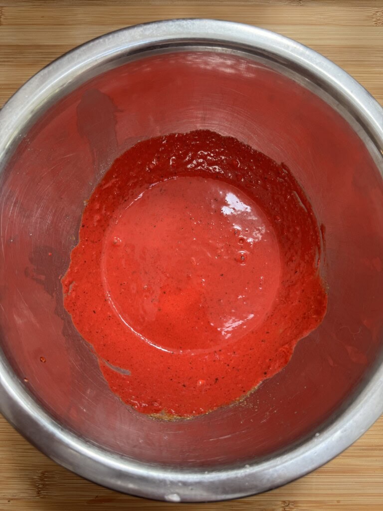A stainless steel mixing bowl containing a bright red liquid mixture, likely a sauce or batter, sits on a wooden surface. The mixture is partially spread around the base and sides of the bowl.