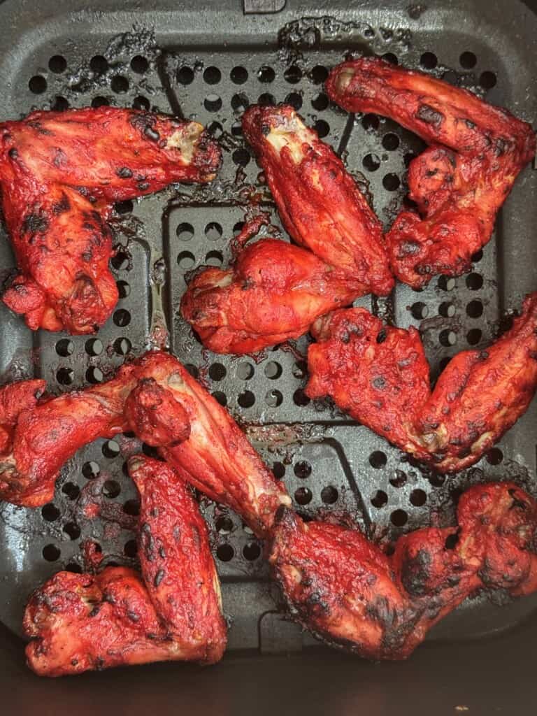 Eight bright red, cooked chicken chaat wings are arranged in a black air fryer basket with visible grill marks and charring on the wings.