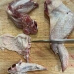 A raw chicken wing is being cut into three parts—drumette, wingette (flat), and tip—on a wooden board with a knife beside the pieces.