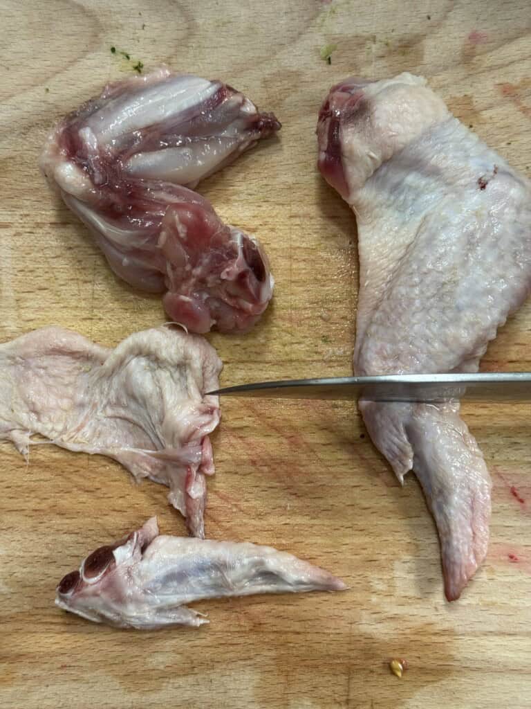 A raw chicken wing is being cut into three parts—drumette, wingette (flat), and tip—on a wooden board with a knife beside the pieces.