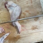 A raw chicken wing on a wooden cutting board is being cut by a large knife. The board shows some food stains and cuts, and part of another chicken wing is visible at the bottom left.