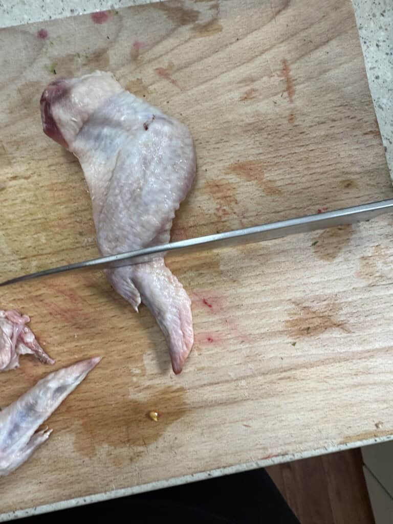 A raw chicken wing on a wooden cutting board is being cut by a large knife. The board shows some food stains and cuts, and part of another chicken wing is visible at the bottom left.