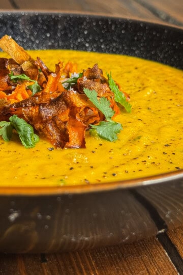 A close-up of a bowl of creamy yellow soup, garnished with crispy vegetable strips and fresh cilantro, served in a black speckled bowl on a wooden table.
