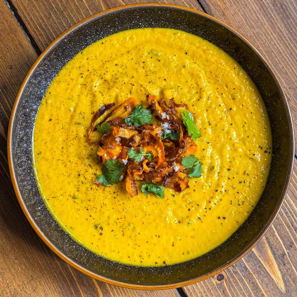 A bowl of golden-yellow soup garnished with crispy fried onions, fresh cilantro leaves, and a sprinkle of black pepper, served on a wooden table.