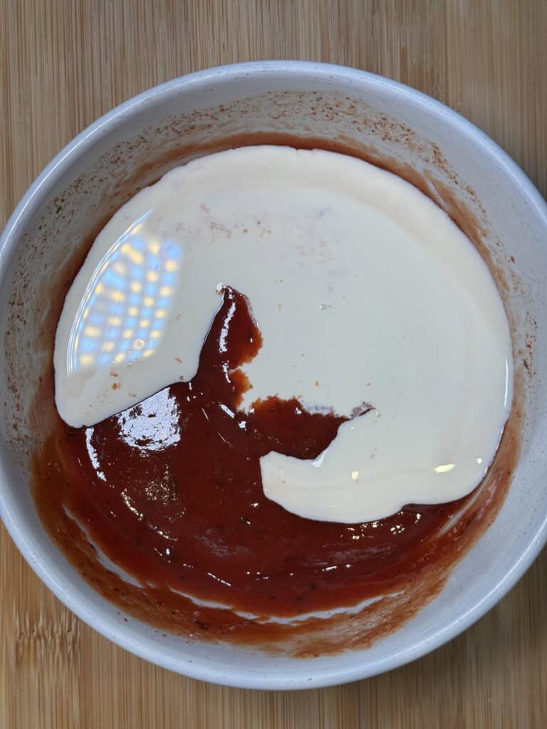 A white bowl on a wooden surface containing a mixture of tomato sauce and cream, with the ingredients partially blended.