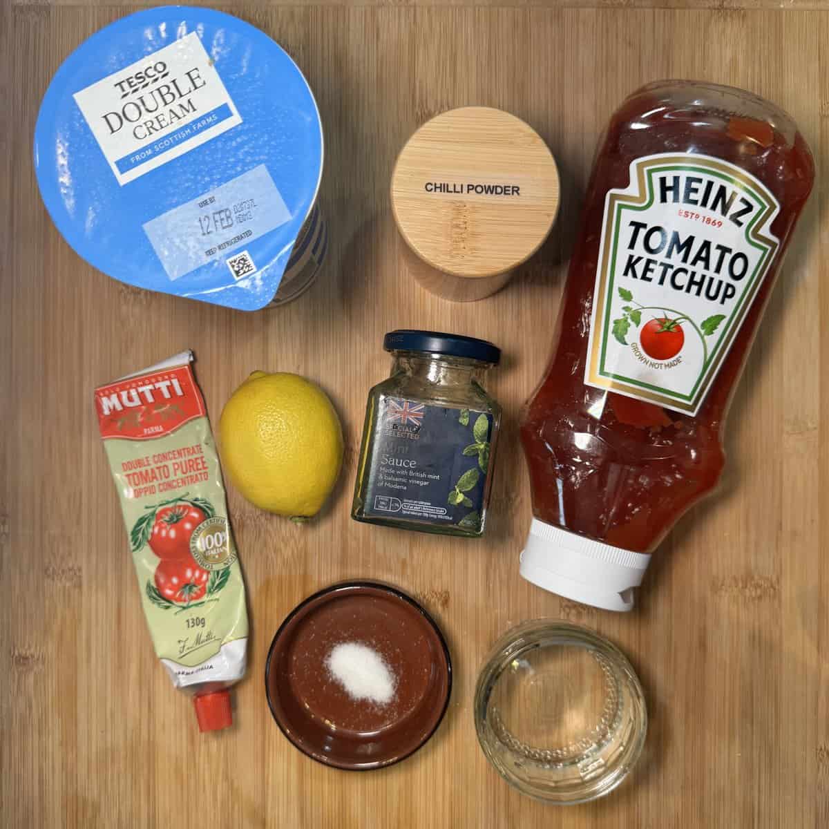 A flat lay of ingredients on a wooden surface: double cream, chilli powder, tomato ketchup, tomato puree, a lemon, dried mixed herbs, a small bowl of salt, and a small bowl of clear liquid.