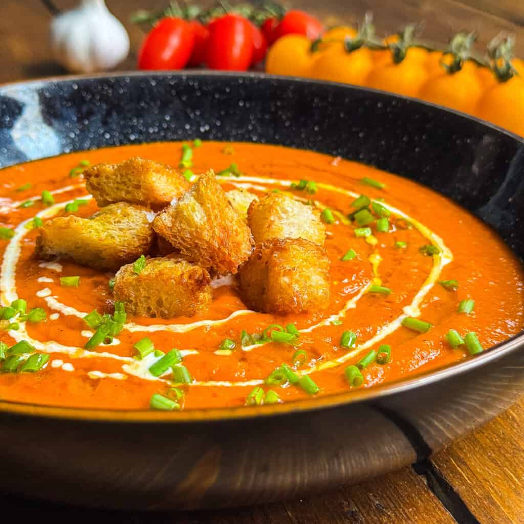 A bowl of creamy tomato soup garnished with a swirl of cream, chopped chives, and golden croutons, with fresh tomatoes and garlic blurred in the background.