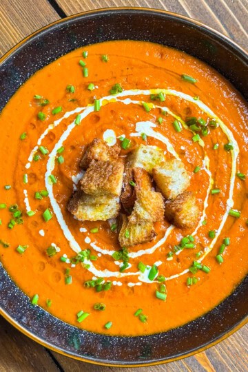A bowl of creamy tomato soup garnished with a swirl of cream, crunchy croutons in the center, and chopped chives, served on a wooden table.