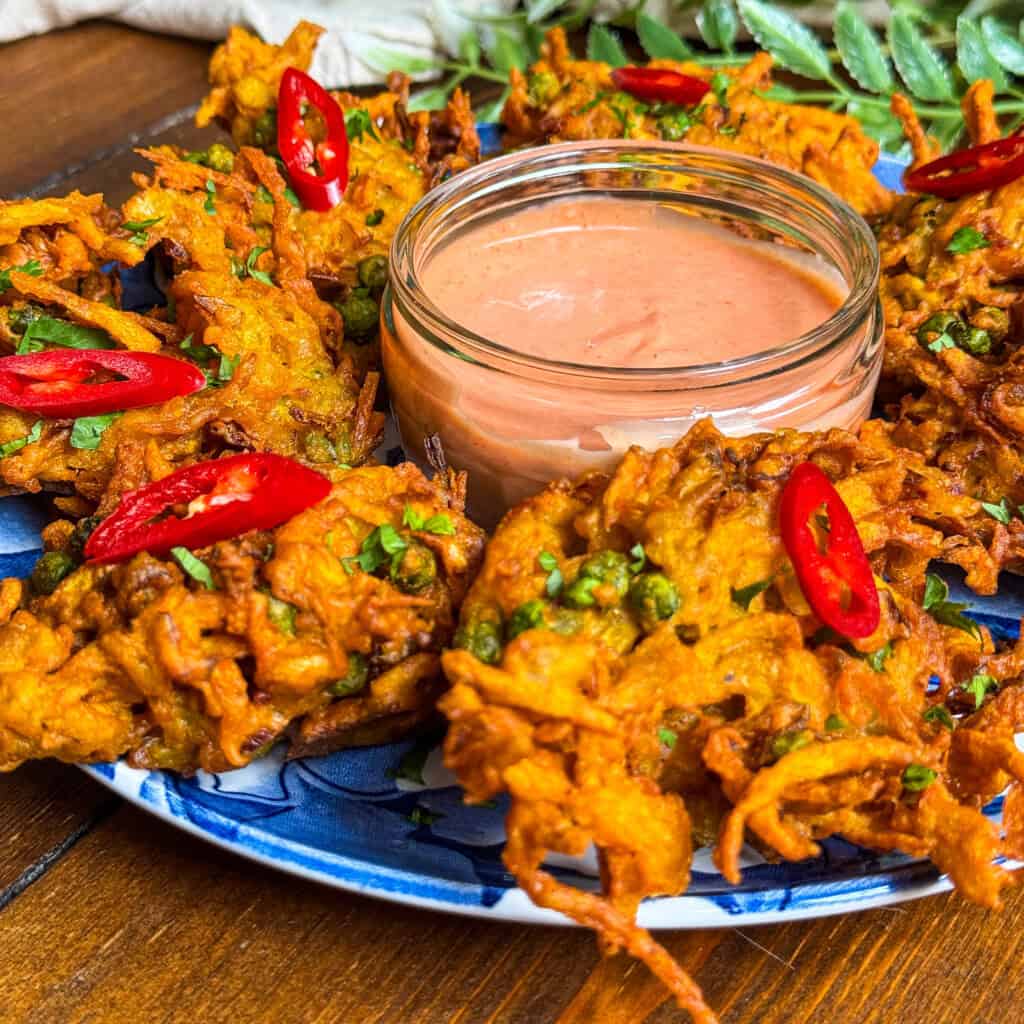 A plate of golden, crispy vegetable bhajis garnished with sliced red chili and herbs, arranged around a small glass bowl of creamy dipping sauce. The dish is vibrant and served on a blue-patterned plate.