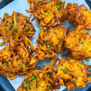 A plate with several golden-brown vegetable bhajis, featuring visible peas and shredded vegetables, sits on a piece of white paper towel to absorb excess oil.