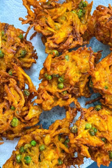 A plate with several golden-brown vegetable bhajis, featuring visible peas and shredded vegetables, sits on a piece of white paper towel to absorb excess oil.