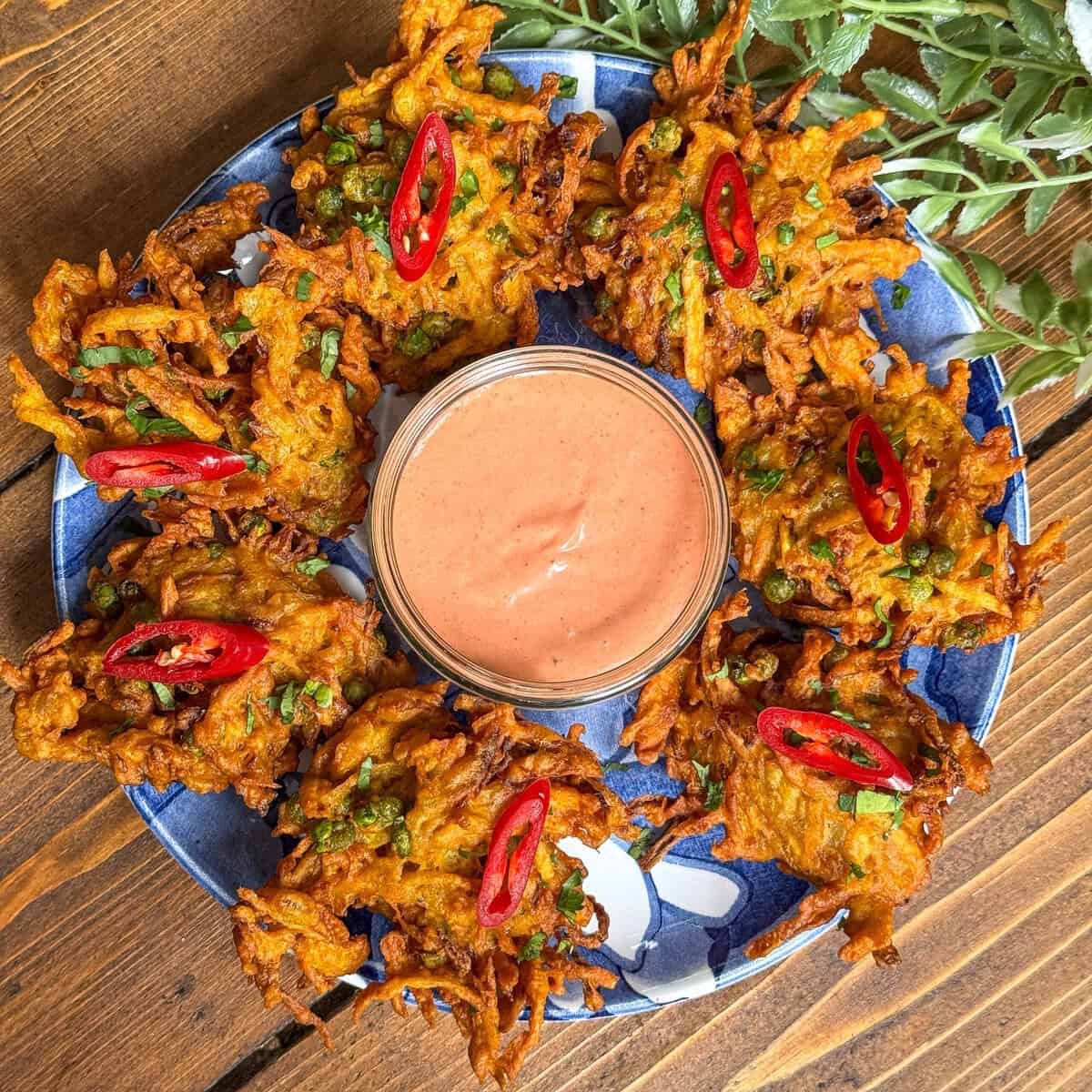A blue plate holds crispy vegetable bhajis garnished with sliced red chili and herbs, arranged in a circle around a bowl of pink dipping sauce. Green leaves are partly visible at the top of the image.