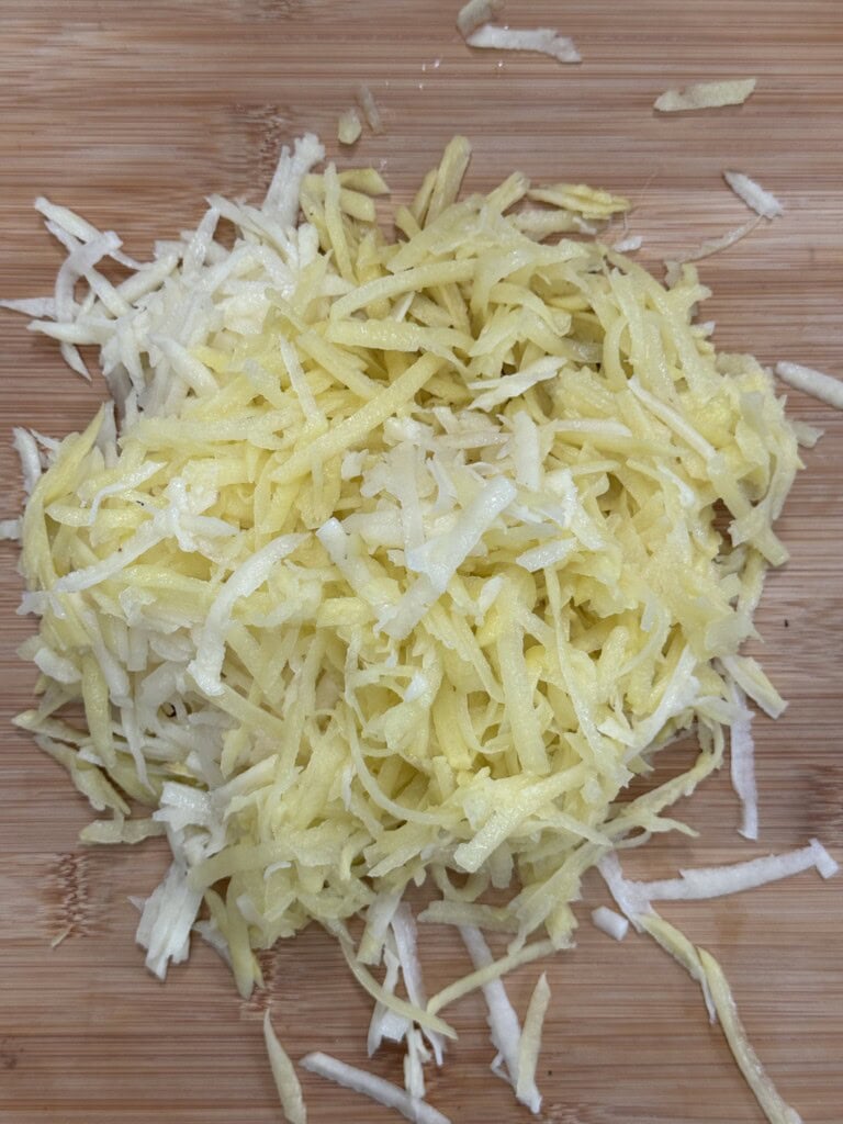 A pile of freshly grated potatoes sits on a wooden cutting board. The potato shreds are pale yellow and white, scattered slightly around the main pile.