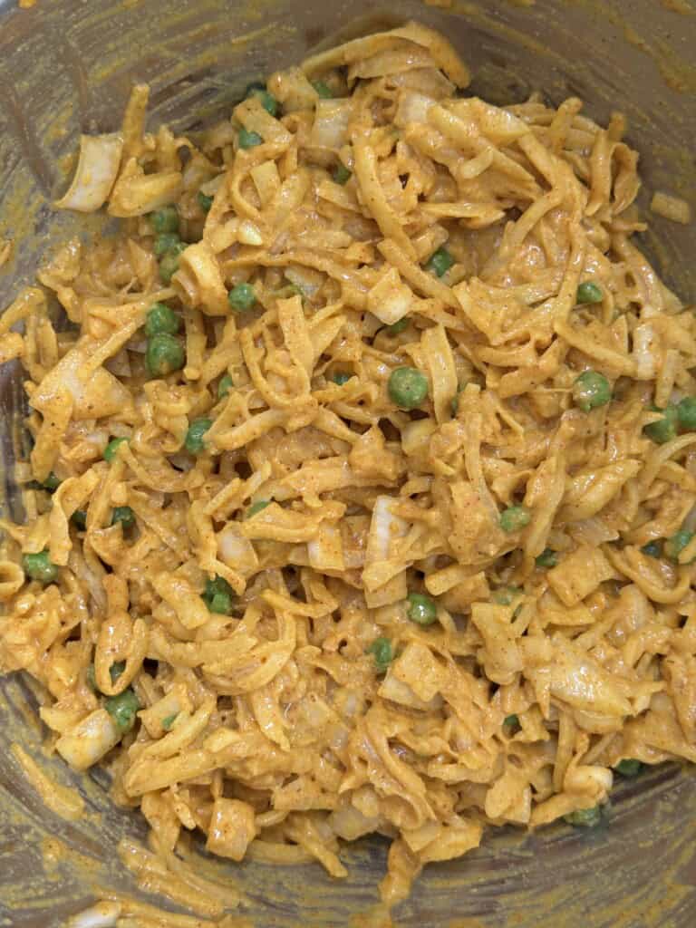 A close-up of a noodle dish mixed with green peas and chopped vegetables, coated in a creamy orange sauce with visible spices.
