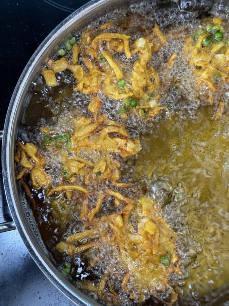 Golden brown fritters with green peas frying in hot oil in a large pan, with bubbles rising around the food, suggesting they are being deep fried.