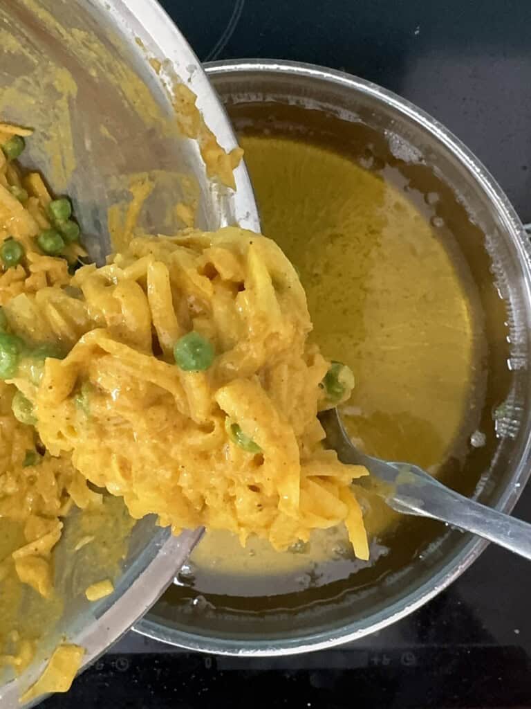 A close-up view of a bowl of creamy yellow pasta with peas being poured into a pot of orange-yellow liquid on a black stovetop.