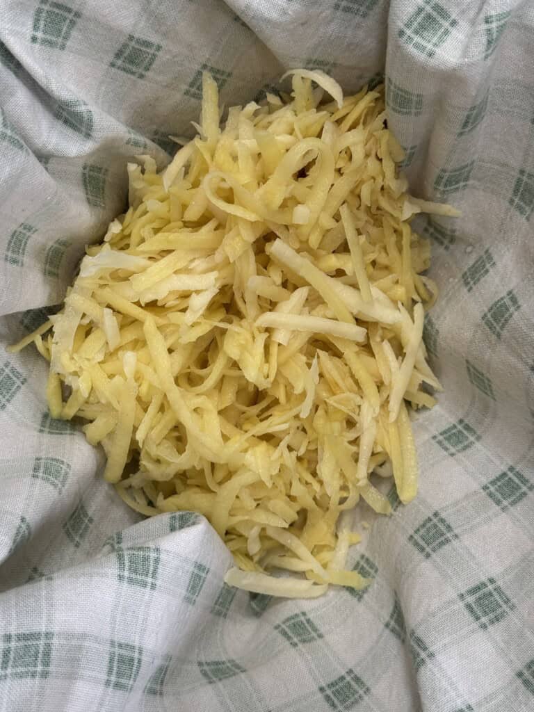 A pile of freshly grated potatoes sits on a white kitchen towel with a green plaid pattern.
