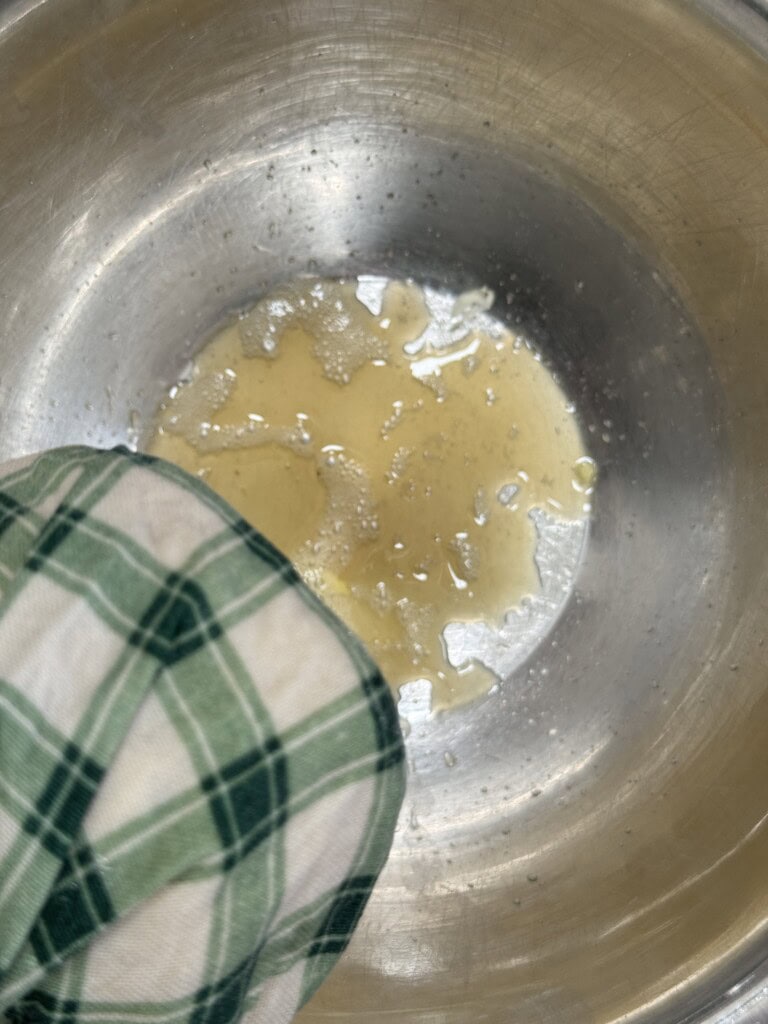 A person wearing a green and white plaid oven mitt holds a metal mixing bowl containing a small amount of beaten eggs.