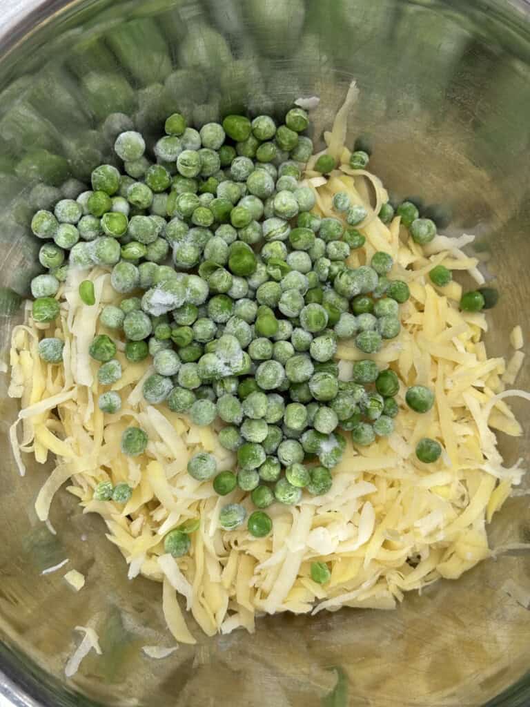 A stainless steel bowl containing a mixture of shredded potatoes and a pile of frozen green peas.
