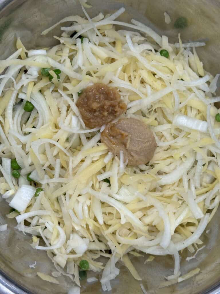A metal bowl containing shredded potatoes, chopped onions, green peas, and two dollops of ginger-garlic paste and brown chutney placed on top.