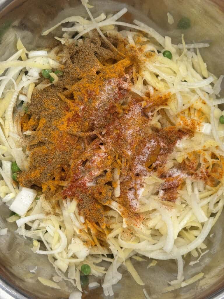 Shredded potatoes, chopped onions, green peas, and various ground spices are mixed in a metal bowl, ready to be combined for cooking.