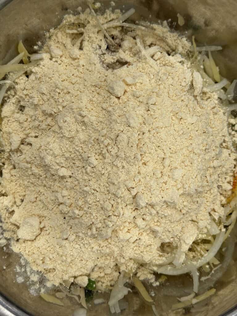 A metal bowl filled with shredded vegetables, partially covered by a thick layer of yellow gram flour (besan) powder.