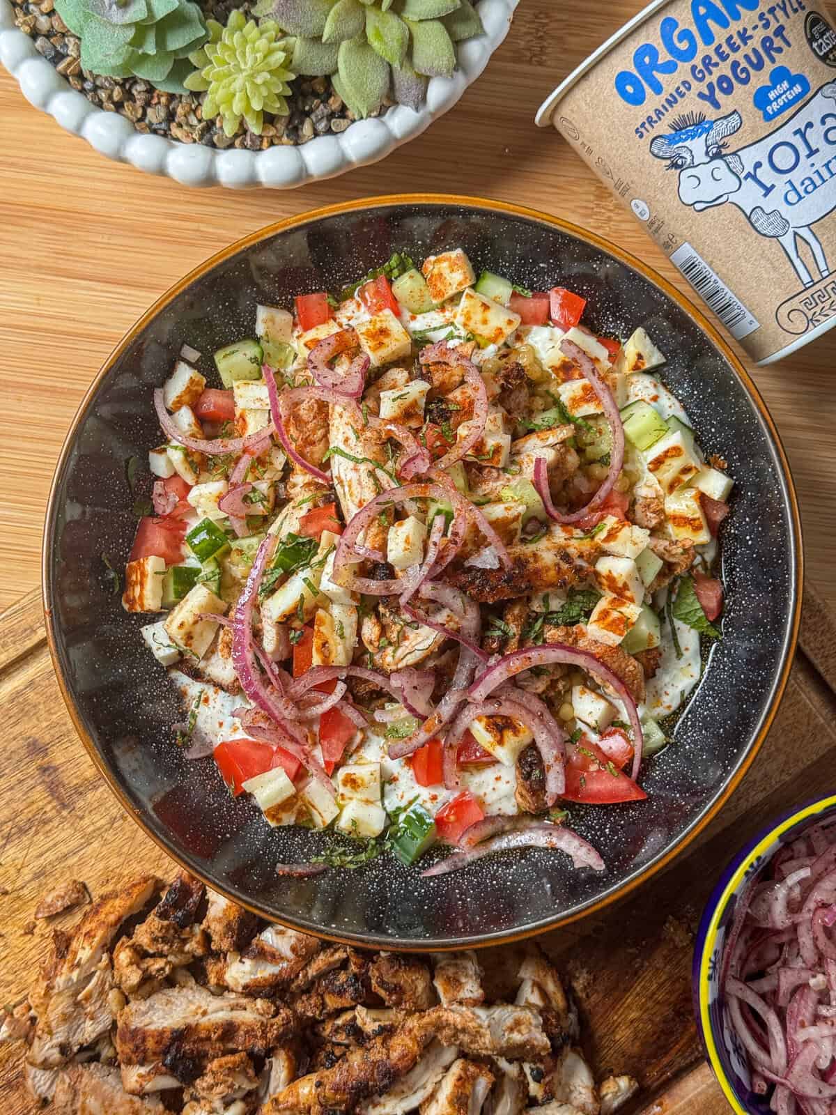A bowl of salad with chopped tomatoes, cucumbers, onions, herbs, and airfryer chicken gyros, garnished with red onion slices. Surrounding items include a yogurt container, succulents, and a wooden board with sliced chicken.